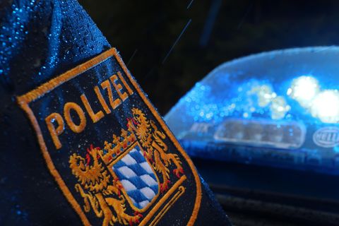 Auf einem regennassen Ärmel prangt das Wappen der Polizei Bayern, während dahinter ein Blaulicht die Dunkelheit erhellt