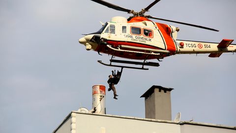 Ein weiß-oranger Hubschrauber fliegt tief über einem Flachdach. Zwischen Dach und Hubschrauber sind die Konturen zweier Menschen