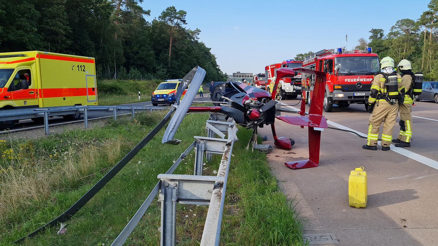 Ein roter Hubschrauber liegt seitlich auf dem Standstreifen einer Autobahn. Die Rotorblätter sind verbogen