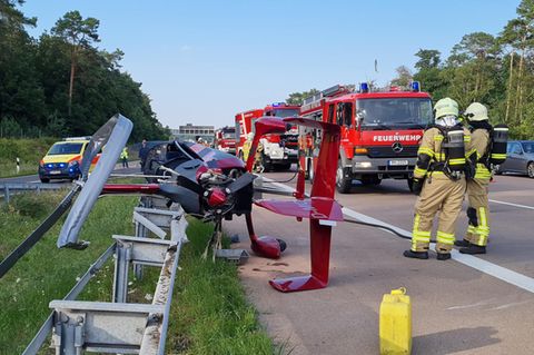 Ein roter Hubschrauber liegt seitlich auf dem Standstreifen einer Autobahn. Die Rotorblätter sind verbogen