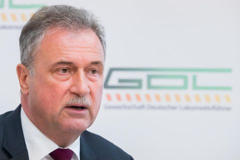 Ein älterer weißer Mann mit grauem Mittelscheitel und Schnauzbart sitzt im Anzug vor einer weißen Wand mit grünem GDL-Logo