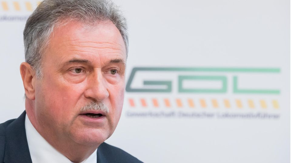 Claus Weselsky GDL Ein älterer weißer Mann mit grauem Mittelscheitel und Schnauzbart sitzt im Anzug vor einer weißen Wand mit grünem GDL-Logo