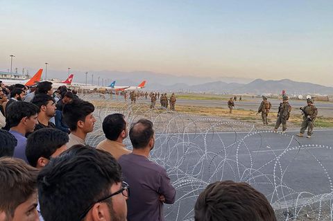 Vor zwei Rollen Nato-Stacheldraht stehen viele dunkelhaarige Männer, dahinter stehen Flugzeuge und Soldaten, die sie bewachen
