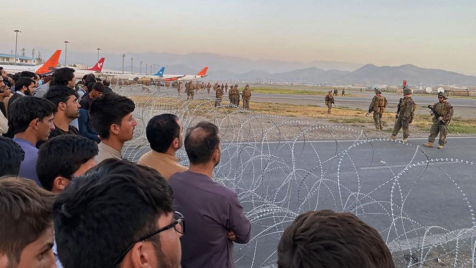 Vor zwei Rollen Nato-Stacheldraht stehen viele dunkelhaarige Männer, dahinter stehen Flugzeuge und Soldaten, die sie bewachen