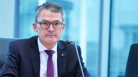 Ein weißer Mann mit angegrautem Seitenscheitel und grau-braunem Brillengestell sitzt in Anzug und Krawatte an einem Holztisch