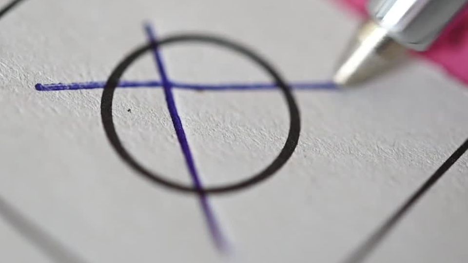 Eine Großaufnahme zeigt einen Kugelschreiber, der ein Kreuz auf einem Wahlzettel malt