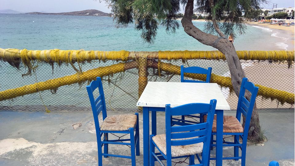 Vier blaue Holzstühle stehen auf einer Strandpromenade um einen Tisch mit weißer quadratischer Platte herum