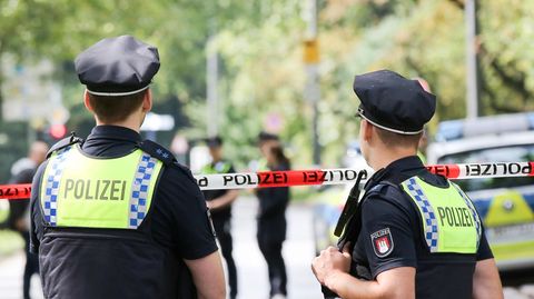 Mit dem Rücken zur Kamera stehen zwei Polizisten in dunkelblauen Uniformen draußen und schauen zu Kollegen hinter Absperrband