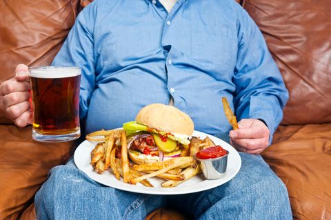 Ungesunde Ernährung: Ein dicker Mann sitzt mit Bier, Burger und Pommes auf dem Sofa