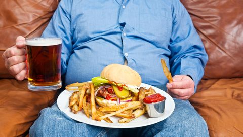 Ungesunde Ernährung: Ein dicker Mann sitzt mit Bier, Burger und Pommes auf dem Sofa