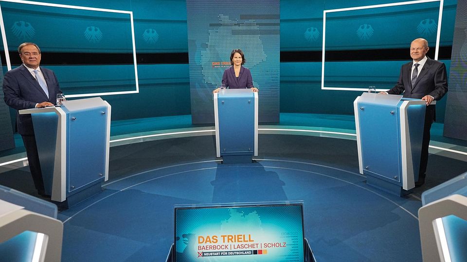 In einem blau beleuchteten TV-Studio stehen zwei Männer und eine Frau an einzelnen Pulten