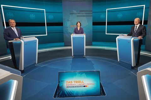 In einem blau beleuchteten TV-Studio stehen zwei Männer und eine Frau an einzelnen Pulten