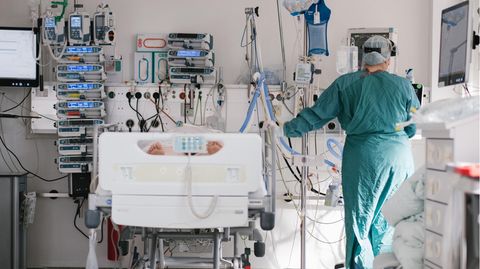 Neben einem Krankenhaus-Bett stehen links zahlreiche Geräte und rechts eine Person in grüner medizinischer Schutzkleidung