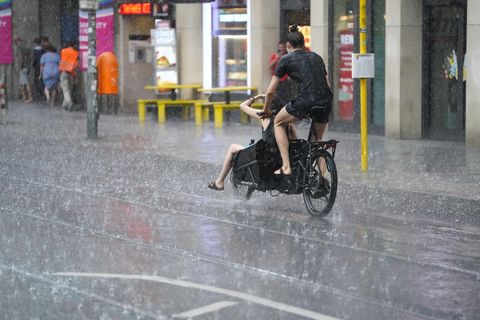 Zwei junge Menschen fahren auf einem Lastenrad - einer auf dem Sattel, einer vorne im Gepäckfach - durch Regen in Berlin