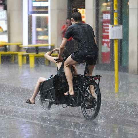 Zwei junge Menschen fahren auf einem Lastenrad - einer auf dem Sattel, einer vorne im Gepäckfach - durch Regen in Berlin