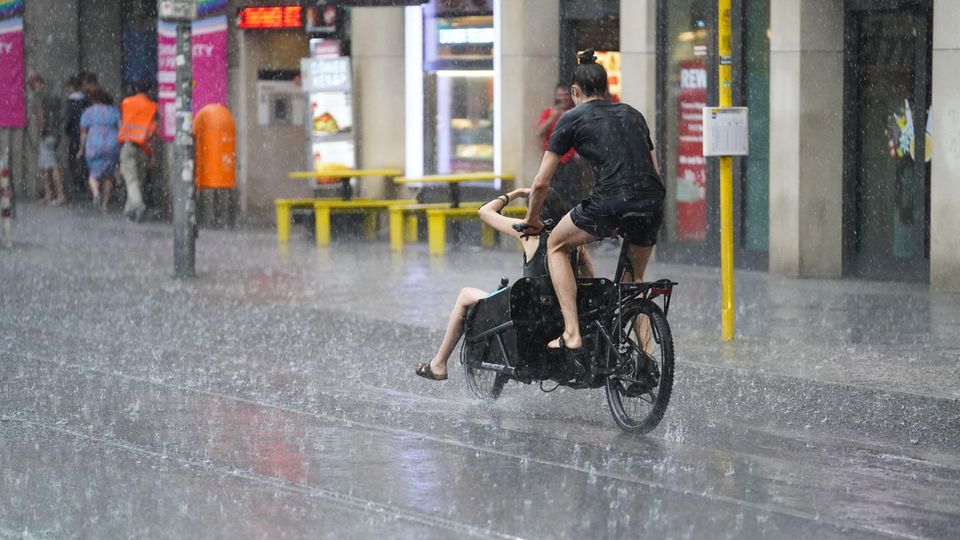Zwei junge Menschen fahren auf einem Lastenrad - einer auf dem Sattel, einer vorne im Gepäckfach - durch Regen in Berlin