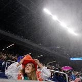 Eine weiße Frau mit langen blonden Haarren zieht sich eine orangene Kapuze über, während sie im Tennisstadion nach oben schaut