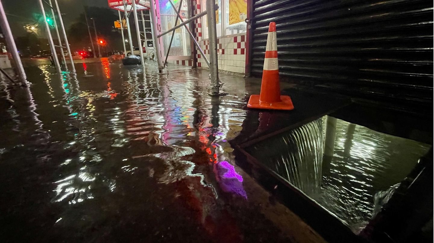Im Dunkeln läuft Wasser durch eine quadratische Öffnung auf dem Fußweg in den Keller eines erleuchteten Fastfood-Restaurants