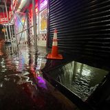 Im Dunkeln läuft Wasser durch eine quadratische Öffnung auf dem Fußweg in den Keller eines erleuchteten Fastfood-Restaurants