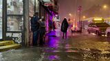 Zwei Männer unterhalten sich vor einem beleuchteten Eck-Laden, während auf überflutetem Fußweg ein Mensch in Regenponcho geht