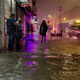 Zwei Männer unterhalten sich vor einem beleuchteten Eck-Laden, während auf überflutetem Fußweg ein Mensch in Regenponcho geht