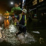 Ein Mann in neongelber Warnweste und mit weißem Sicherheitshelm stapft durch knöcheltiefes Wasser auf einer dunklen Straße
