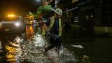 Ein Mann in neongelber Warnweste und mit weißem Sicherheitshelm stapft durch knöcheltiefes Wasser auf einer dunklen Straße