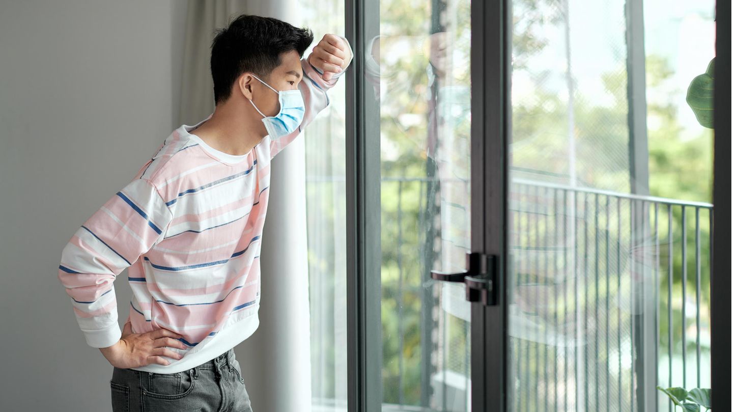 Young Vietnamese stands with mask in front of the window and looks longingly outside.