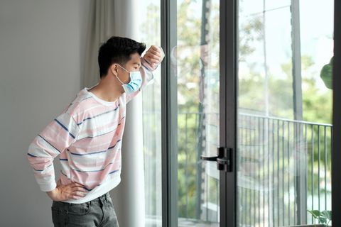 Junger Vietnamese steht mit Maske vorm Fenster und schaut sehnsüchtig nach draußen.