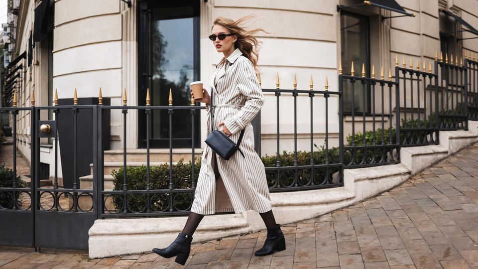Frau läuft mit Kaffee in der Hand die Straße entlang und trägt einen Trenchcoat