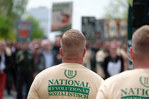 Zwei weiße, junge Männer mit blonden Haaren und ausrasierten Nacken tragen beige T-Shirts mit dem Logo von "Der dritte Weg"