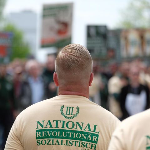 Zwei weiße, junge Männer mit blonden Haaren und ausrasierten Nacken tragen beige T-Shirts mit dem Logo von "Der dritte Weg"