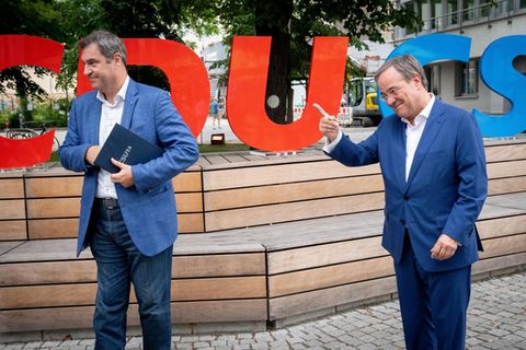 Auf hölzernen Stufen stehen die roten Buchstaben CDU und die blauen Buchstaben CSU, davor zwei weiße Männer in bblauen Anzügen