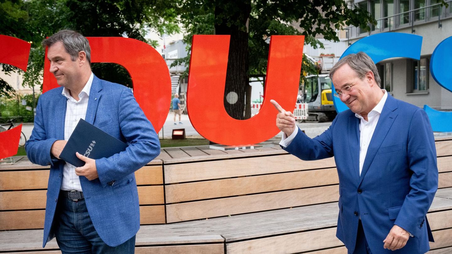 Auf hölzernen Stufen stehen die roten Buchstaben CDU und die blauen Buchstaben CSU, davor zwei weiße Männer in bblauen Anzügen