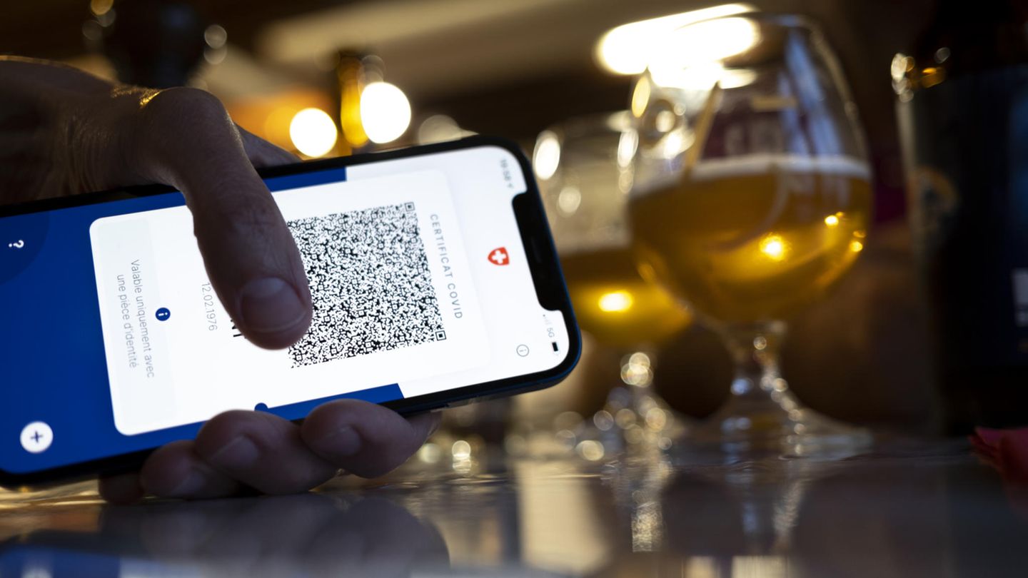Person hält ein Smartphone mit Corona-Zertifikat, im Hintergrund zwei Biergläser
