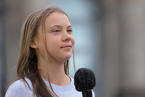 Eine junge weiße Frau mit langen, dunkelblonden Haaren lächelt leicht, während sie ein schwarzes Mikro hält