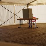 In einem Festzelt mit Holzboden steht eine Wahlkabine, in der gerade ein Mensch steht und wählt