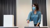 Eine weiße Frau mit schwarz-braunen schulterlangen Haaren und blauer Bluse wirft einen Zettel in ein Wahlurne