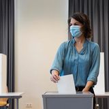 Eine weiße Frau mit schwarz-braunen schulterlangen Haaren und blauer Bluse wirft einen Zettel in ein Wahlurne