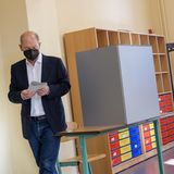 Ein weißer Mann mit Halbglatze, dunkelblauem Jackett und Jeans faltet beim Gehen aus der Wahlkabine einen Zettel