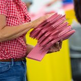 Eine weiße Frau mit rot-weiß karierter Bluse trägt einen Stapel roter Briefwahl-Umschläge mit beiden Händen