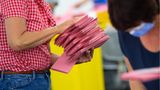 Eine weiße Frau mit rot-weiß karierter Bluse trägt einen Stapel roter Briefwahl-Umschläge mit beiden Händen