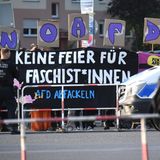 Hinter von Polizisten und rot-weißen Absperrgittern stehen Protestierende mit einem Banner "Keine Feier für Faschist*innen"