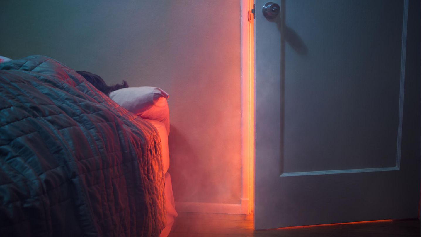 Frau schläft im Bett. Durch die offene Zimmertür dringt Rauch und orangenes Licht ein.