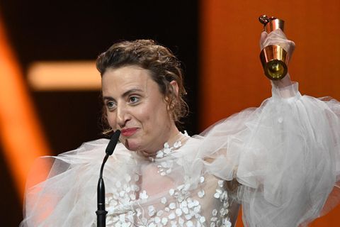 Eine dunkelblonde weiße Frau regt ihren linken Arm mit einer goldenen Trophäe hoch. Sie trägt ein halbtransparentes weißes Kleid