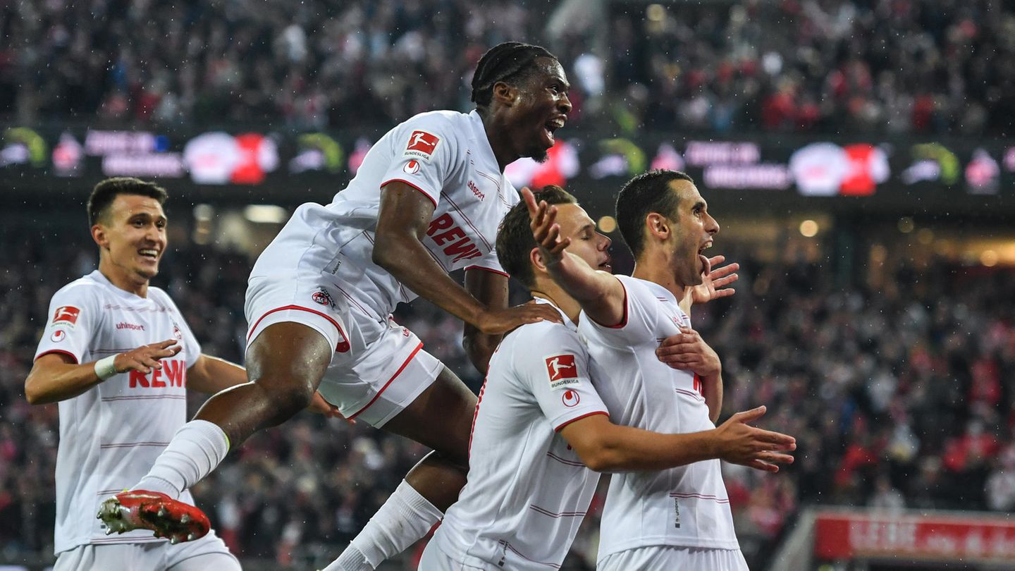 Eine Horde Fußballer in weißen Trikots und Hosen jubelt hinter einem Mitspieler, der seine Arme ausbreitet