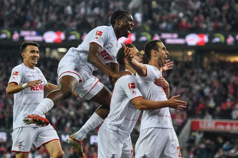 Eine Horde Fußballer in weißen Trikots und Hosen jubelt hinter einem Mitspieler, der seine Arme ausbreitet