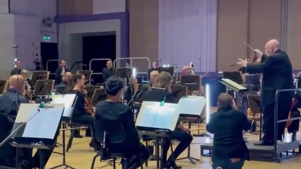 Trotz Demenz: Vater dirigiert ungeachtet seiner Krankheit Orchester Ein Orchester sitzt im Halbkreis um ein Dirigentenpult. Auf dem Pult steht ein Senior, der mit ausgestreckten Armen dirigiert.