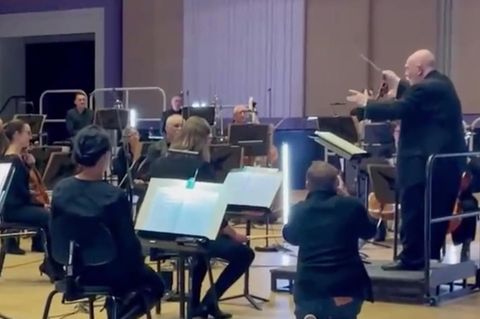 Ein Orchester sitzt im Halbkreis um ein Dirigentenpult. Auf dem Pult steht ein Senior, der mit ausgestreckten Armen dirigiert.