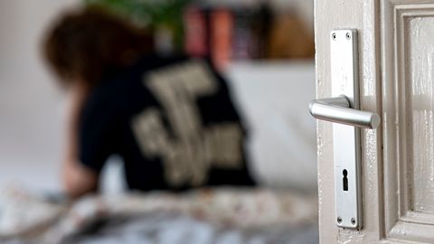 Junge Frau sitzt mit Rücken zur Kamera und gesenktem Kopf auf dem Bett